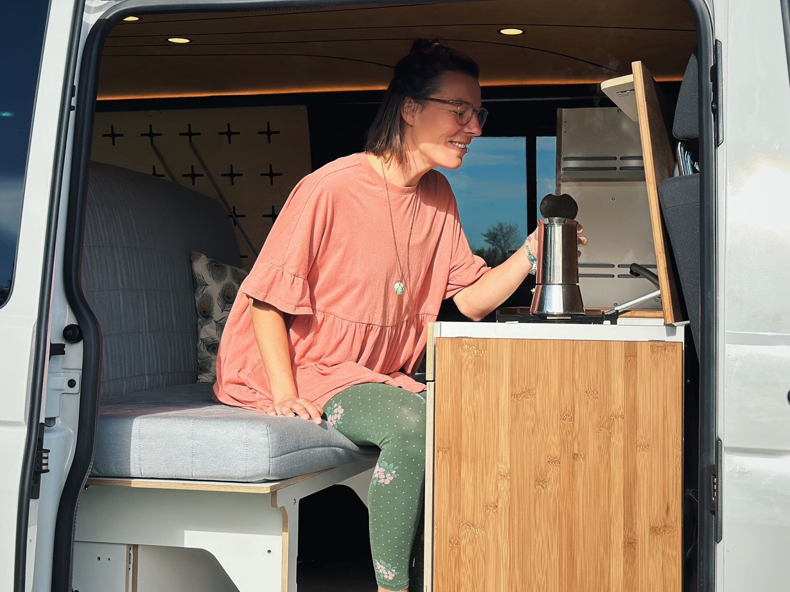 Osoba sedí v karavanu a připravuje kávu u kuchyňského modulu.