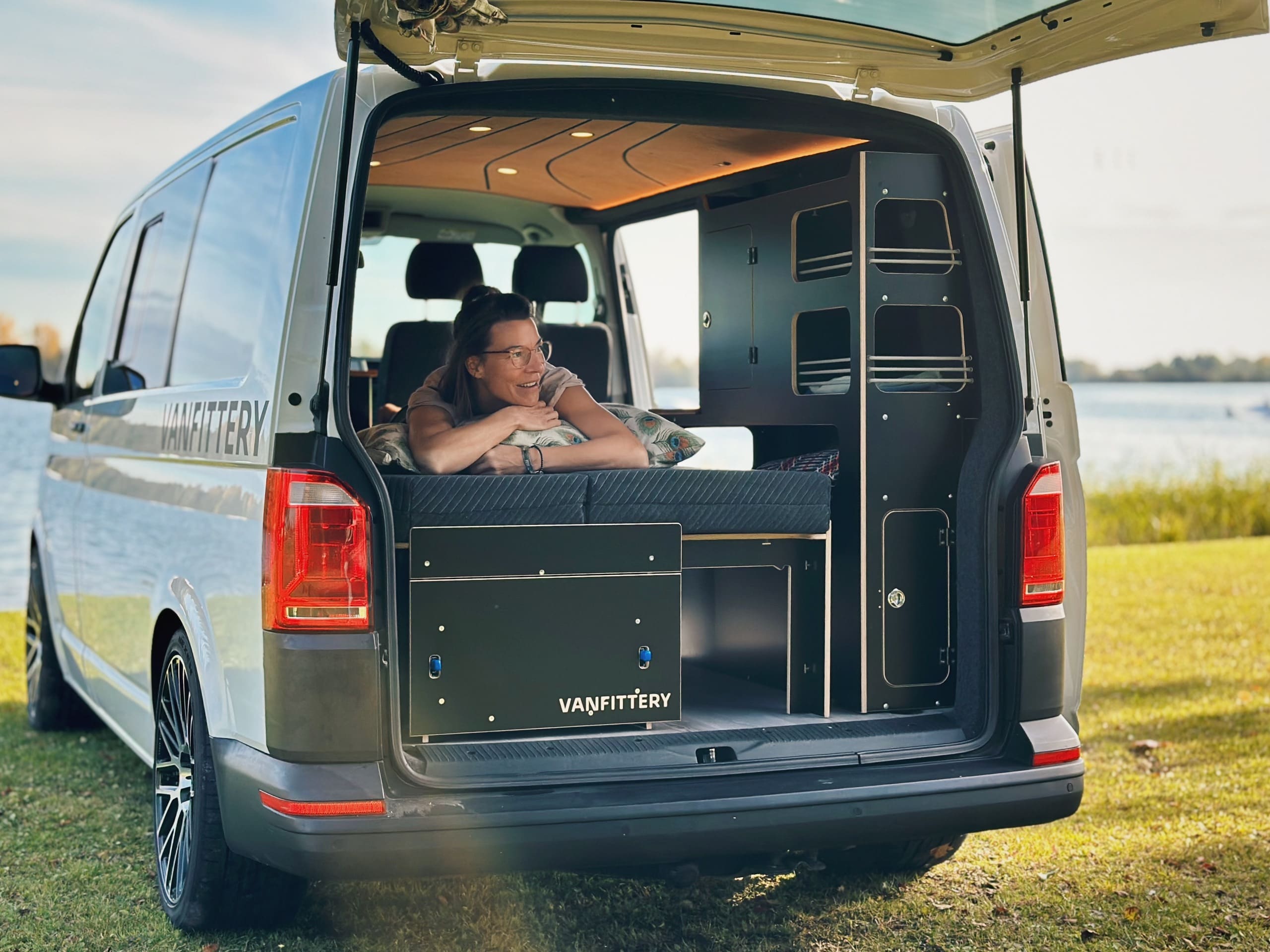 Eine Frau entspannt sich in einem umgebauten Van am Wasser, während sie auf einem Camping-Bettsystem liegt.