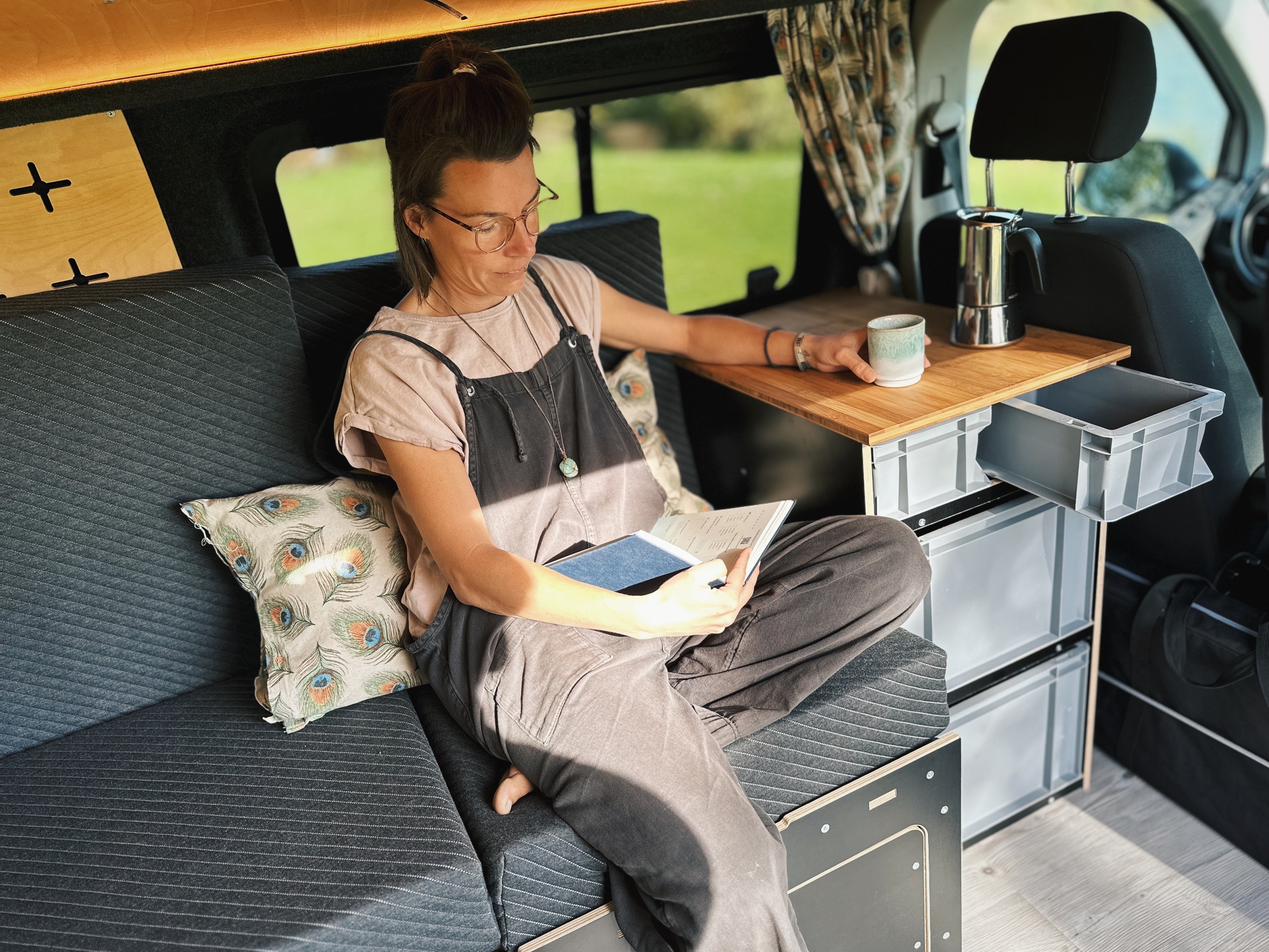 V obytném voze sedí člověk uvolněně na pohodlném sedadle, čte si knihu a vychutnává si kávu, zatímco vedle něj se nachází praktická kuchyňka s úložným prostorem a stolem.