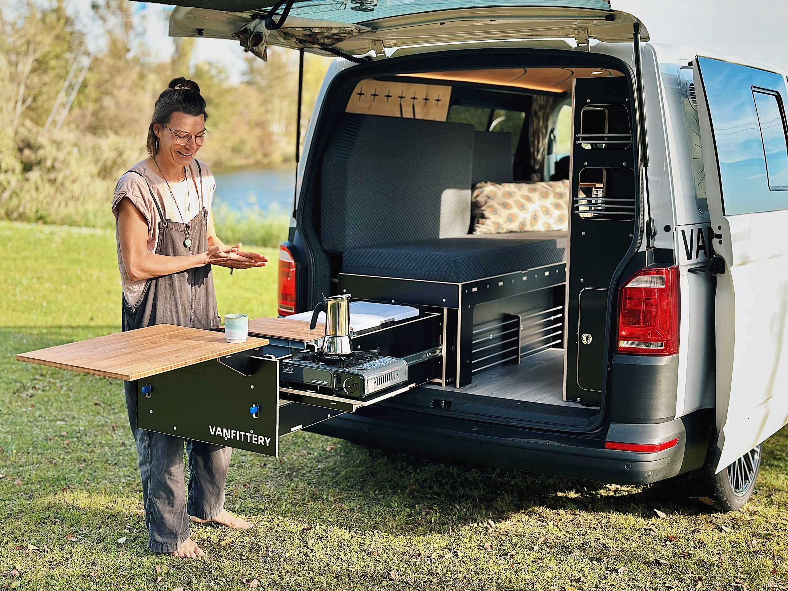 Una persona prepara una comida en el portón trasero abierto de una autocaravana mientras trabaja en un módulo de cocina extraíble.