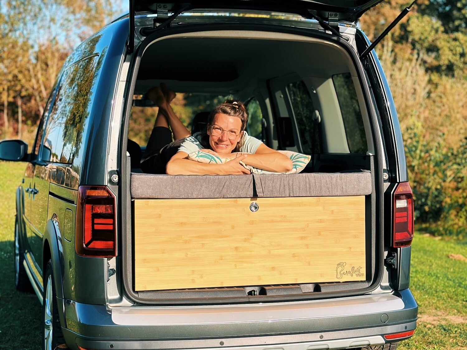 Jedna osoba relaksuje się w otwartej tylnej części pojazdu kempingowego, który jest wyposażony w przytulną część sypialną otoczoną naturalnym krajobrazem.