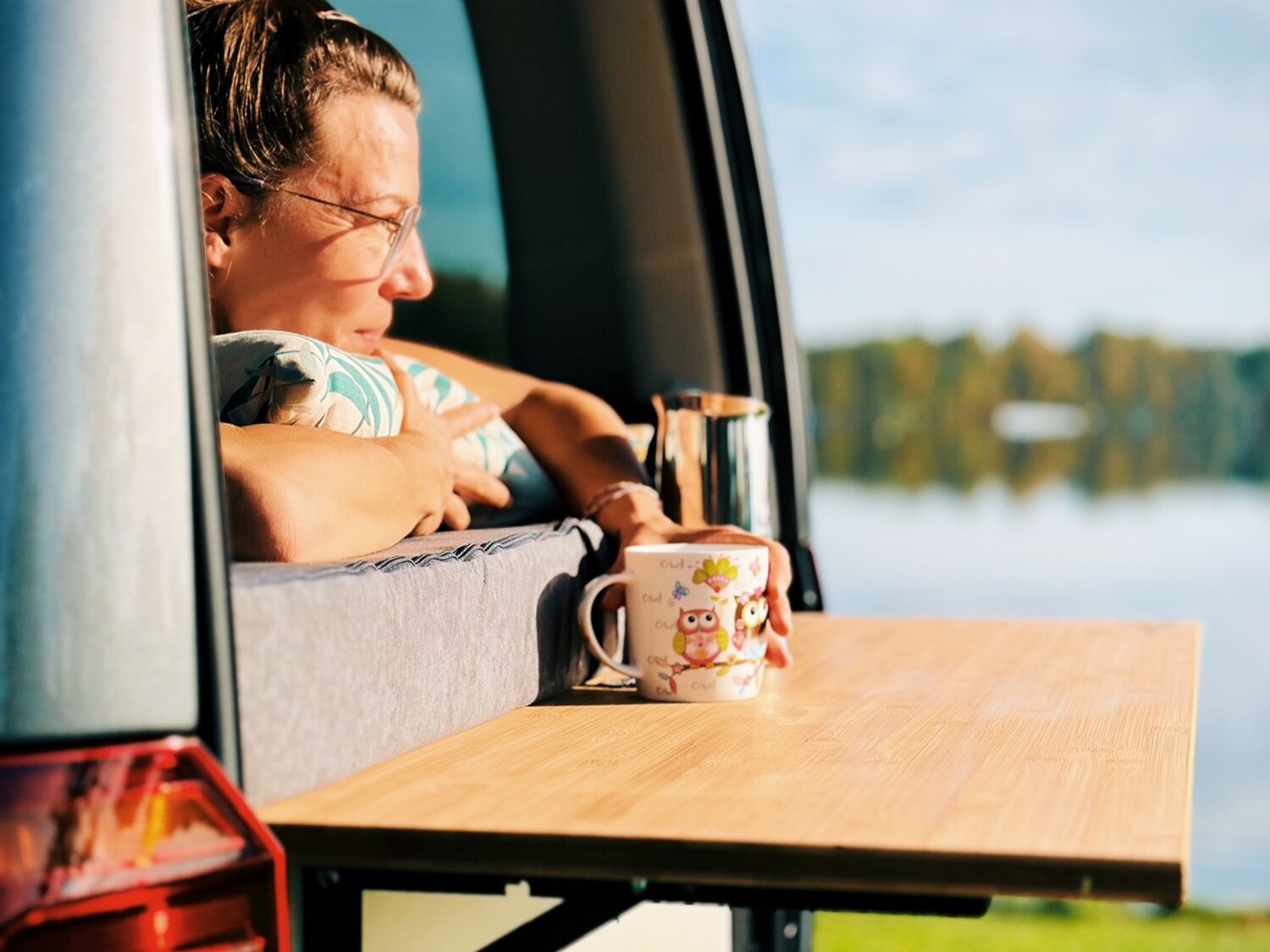 Osoba relaksuje się przy otwartym oknie furgonetki, trzymając poduszkę i delektując się filiżanką kawy na składanym stoliku z widokiem na spokojne jezioro.