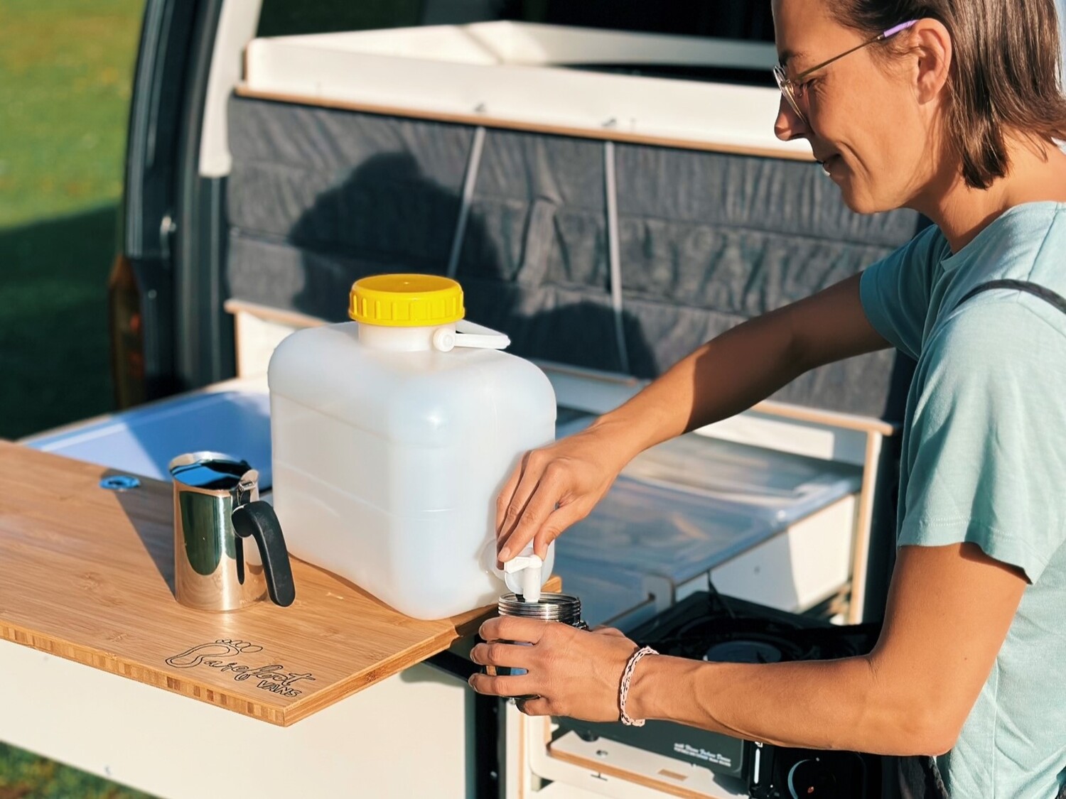 Osoba nalewa wodę z kanistra do pojemnika na kempingowym stole kuchennym, podczas gdy w tle widać mobilną kuchnię.
