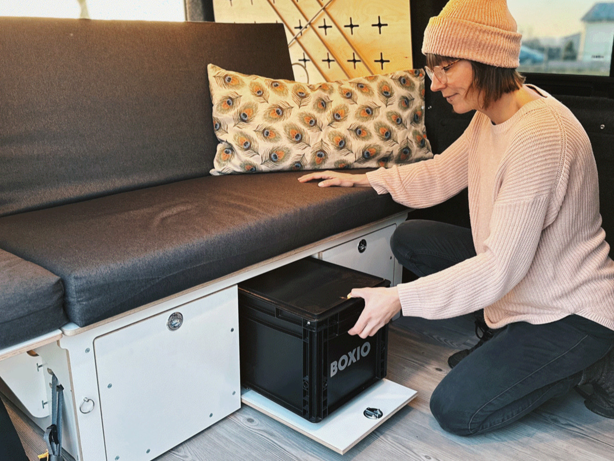 A person sits on a sofa and pulls a camping box out of a storage space to demonstrate the practical use of camping furniture.