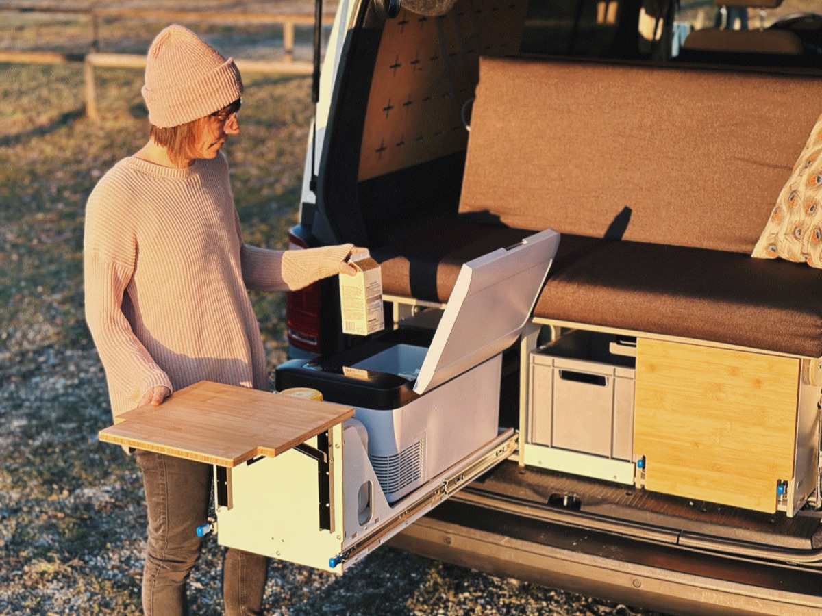 A person in a light-coloured jumper and cap stands next to an open van and uses a pull-out wooden plate while holding a carton of milk in his hand.
