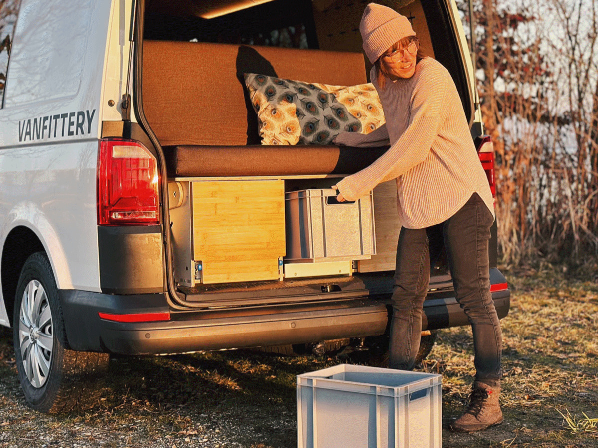 One person takes a box out of the boot of a camper van, which is equipped with cosy furniture and a decorative cushion.