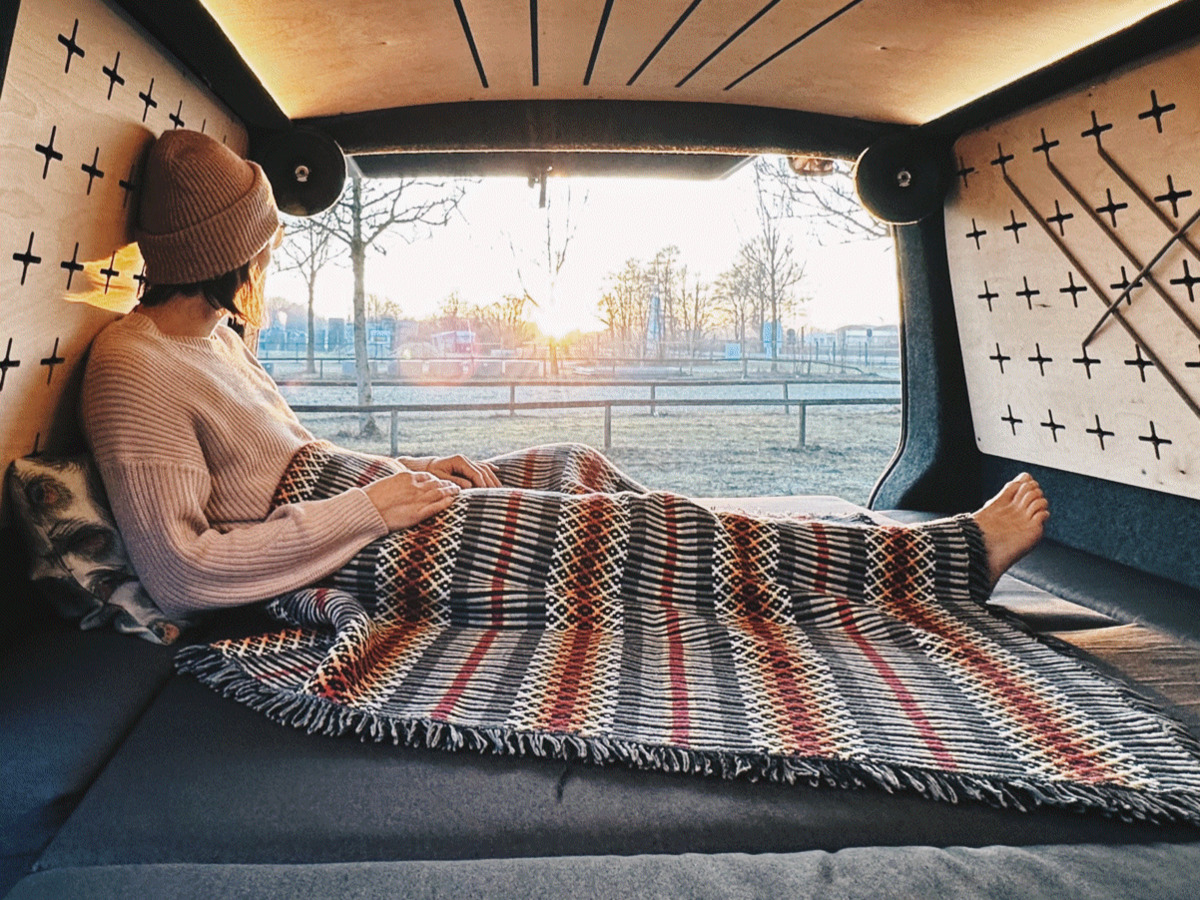A person sits relaxed in a camper van and enjoys the sunset while wrapped in a colourful blanket.