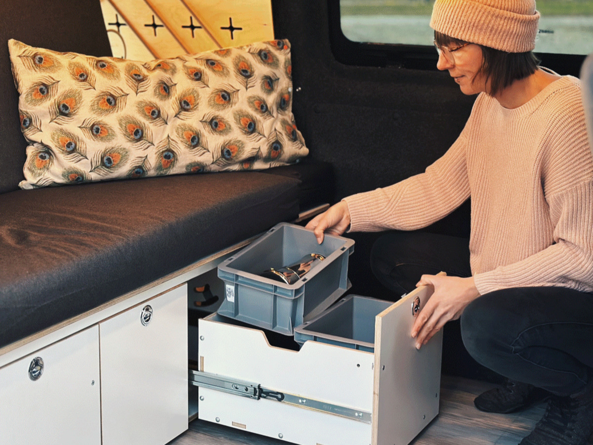 One person sits in a camper and organises camping accessories in a practical cupboard module with drawers.
