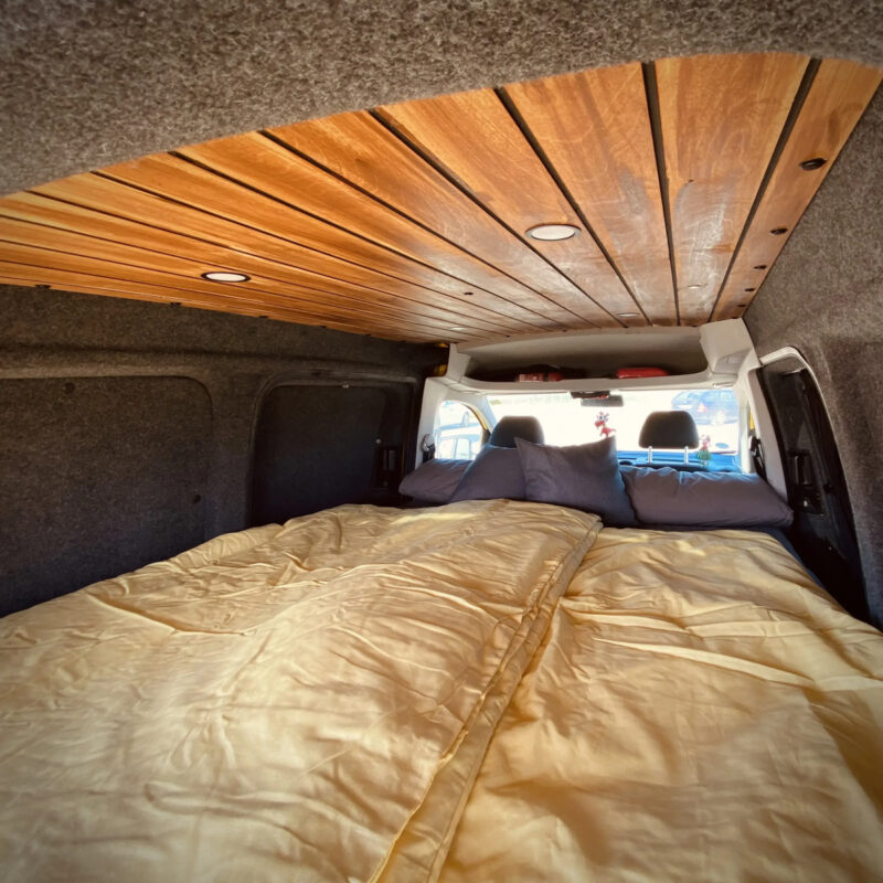 Útulný spací systém v kempinkovém vozidle s dřevěným obložením, vybavený dekou a polštářem pro pohodlný spánek.