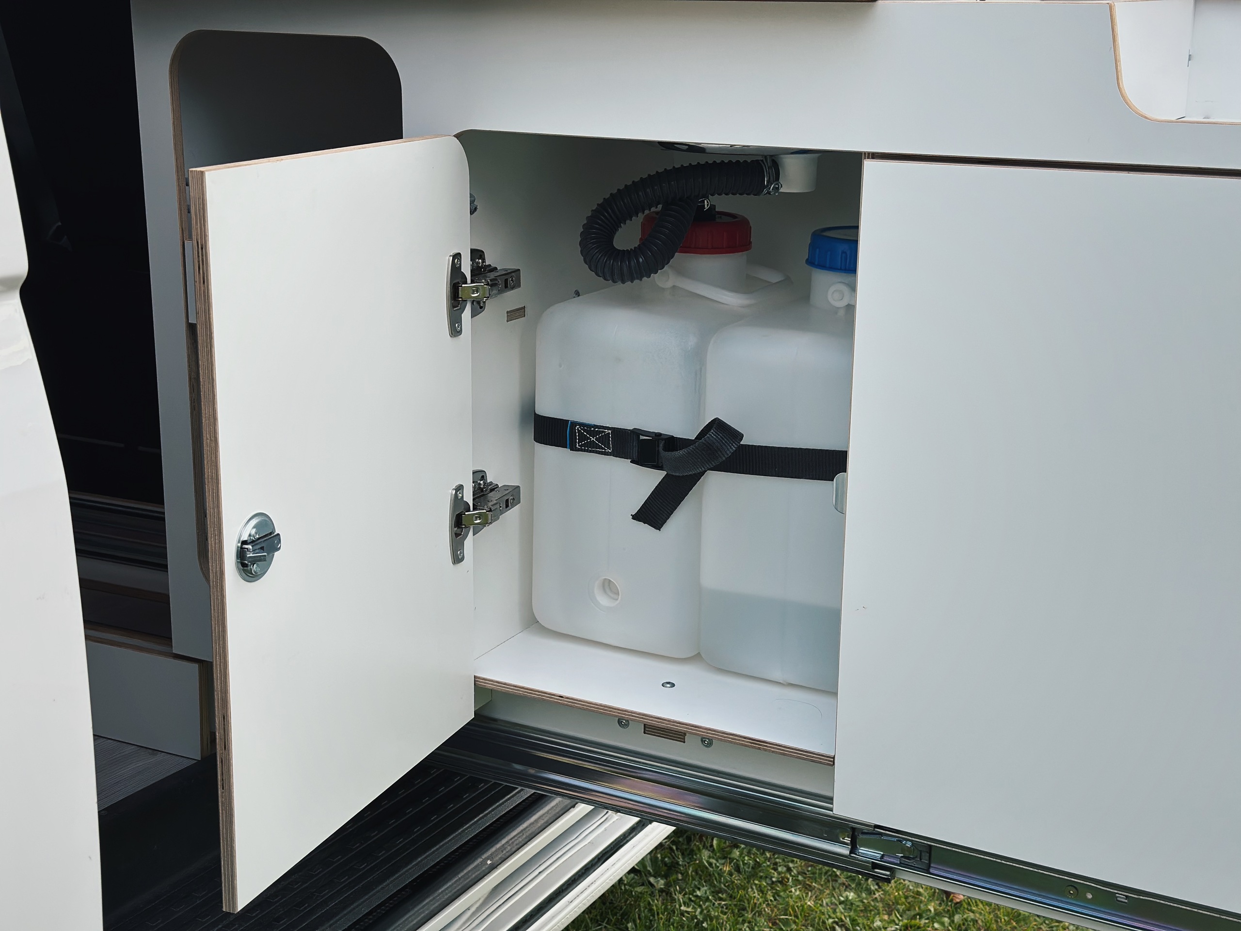 Otevřená skříňka v obytném vozidle se dvěma nádržemi na vodu bezpečně připevněnými popruhem.