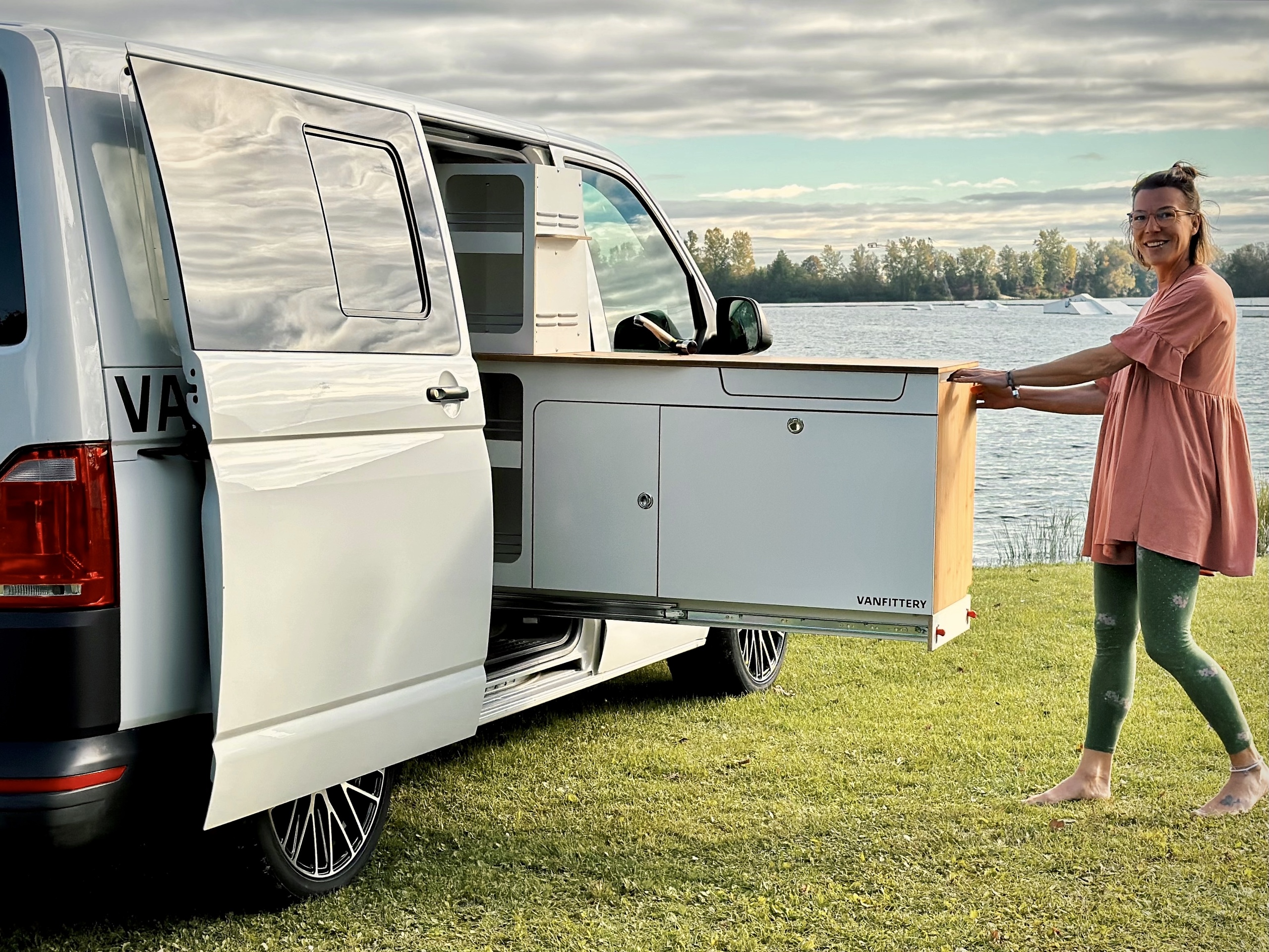 Jedna osoba připravuje kuchyňku z obytného vozu pod širým nebem, obklopená malebnou krajinou u vody.
