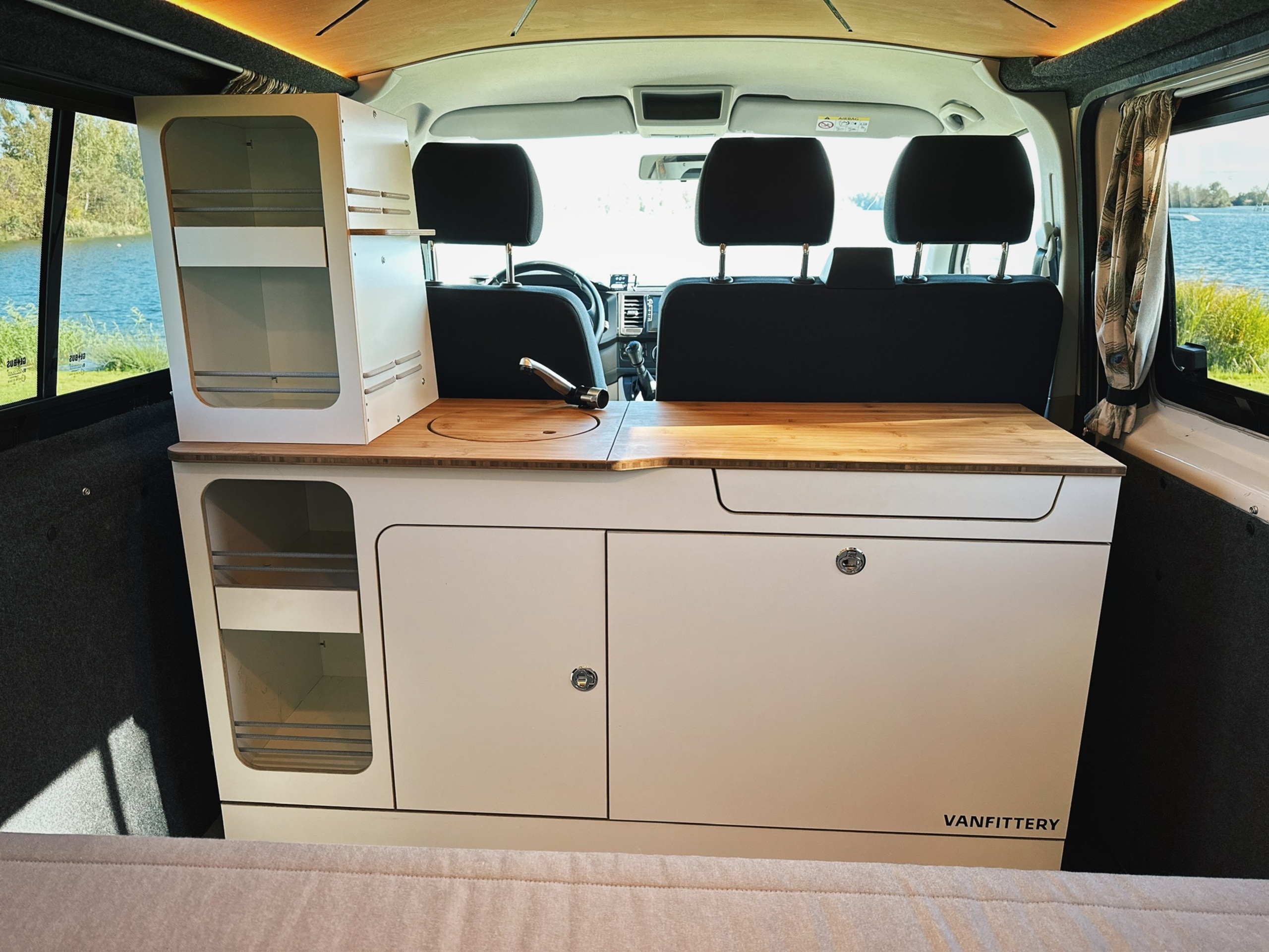 Moderní kempinková kuchyně v interiéru dodávky s dřevěnou pracovní deskou, úložným prostorem a výhledem na jezero v pozadí.