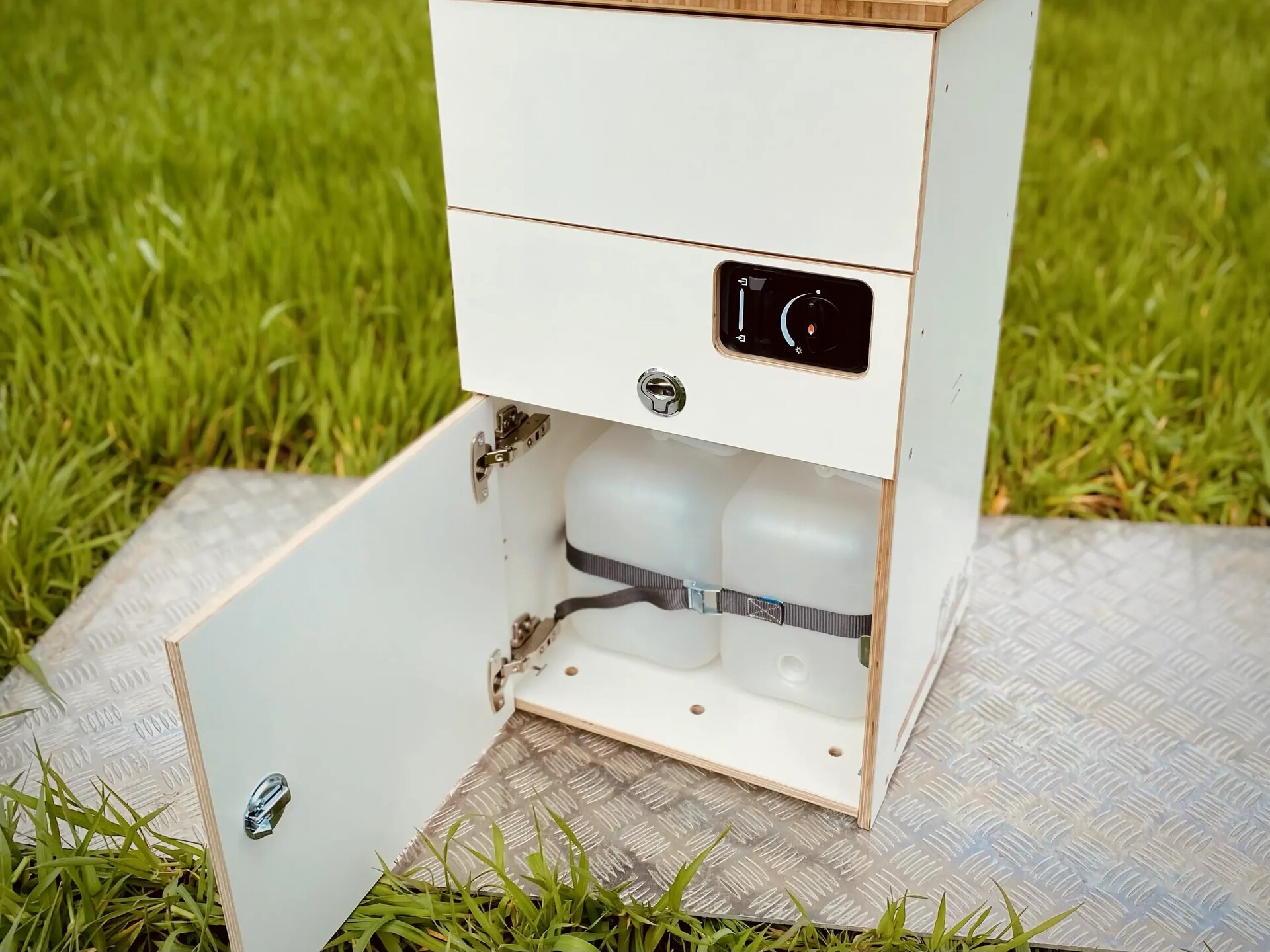 Et moderne campingskab med to skuffer og et rum til vandbeholdere, placeret på en stabil base omgivet af grønt græs.