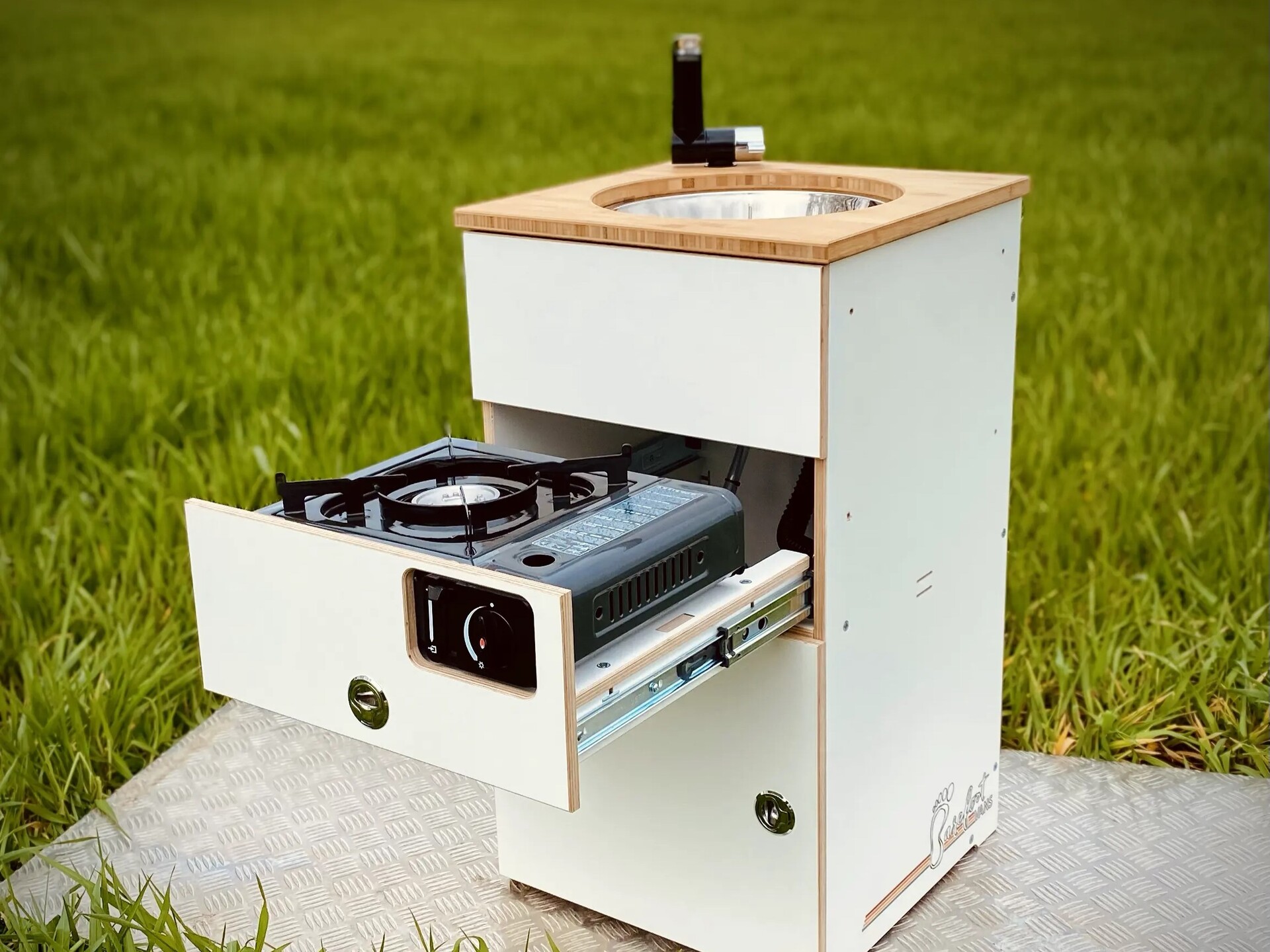 Et kompakt campingkøkkenmodul med gaskomfur og træbordplade, placeret på en eng.