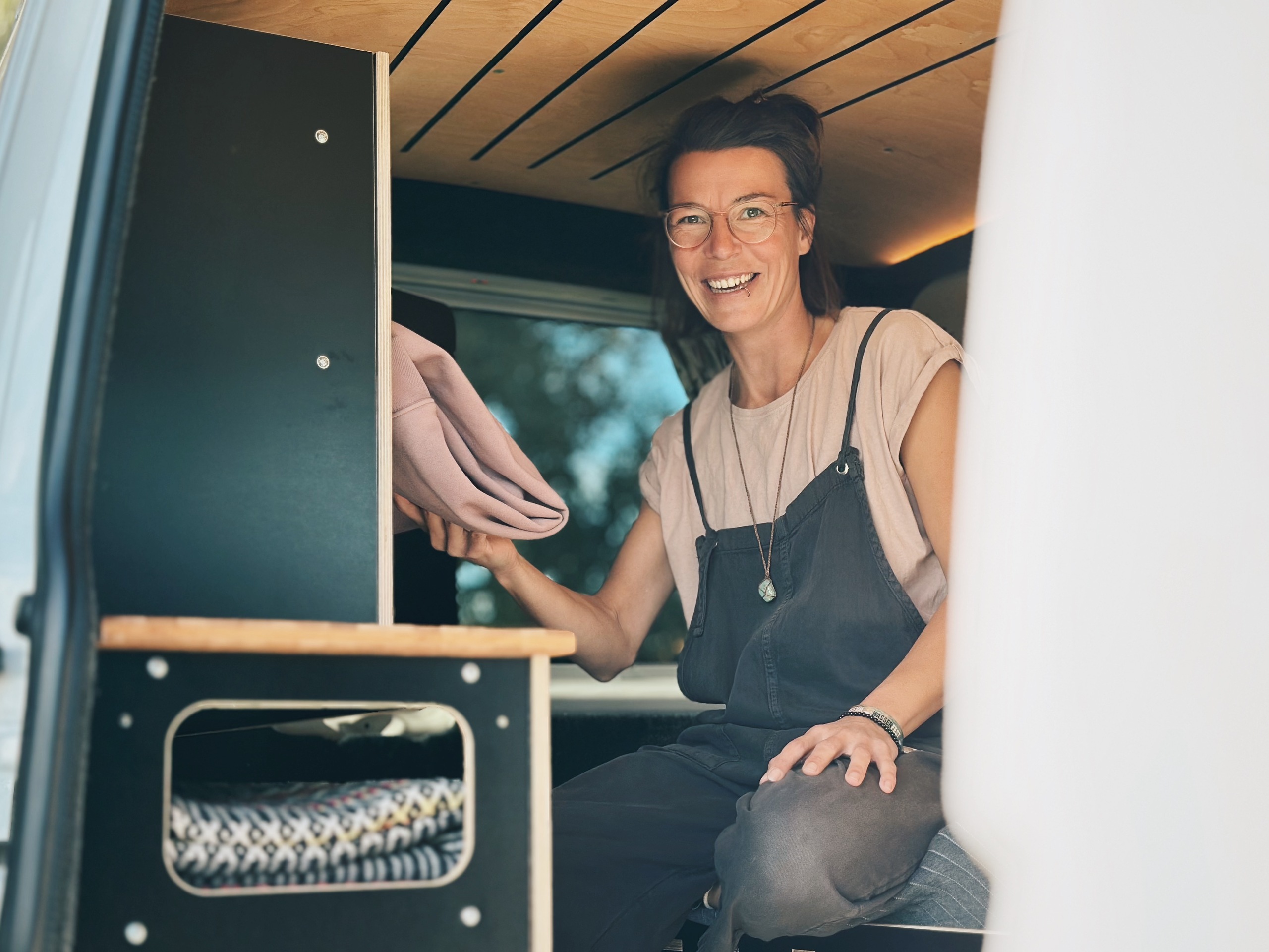 Usměvavá žena sedí v obytném voze se složenou dekou, obklopená kempingovým nábytkem a doplňky.