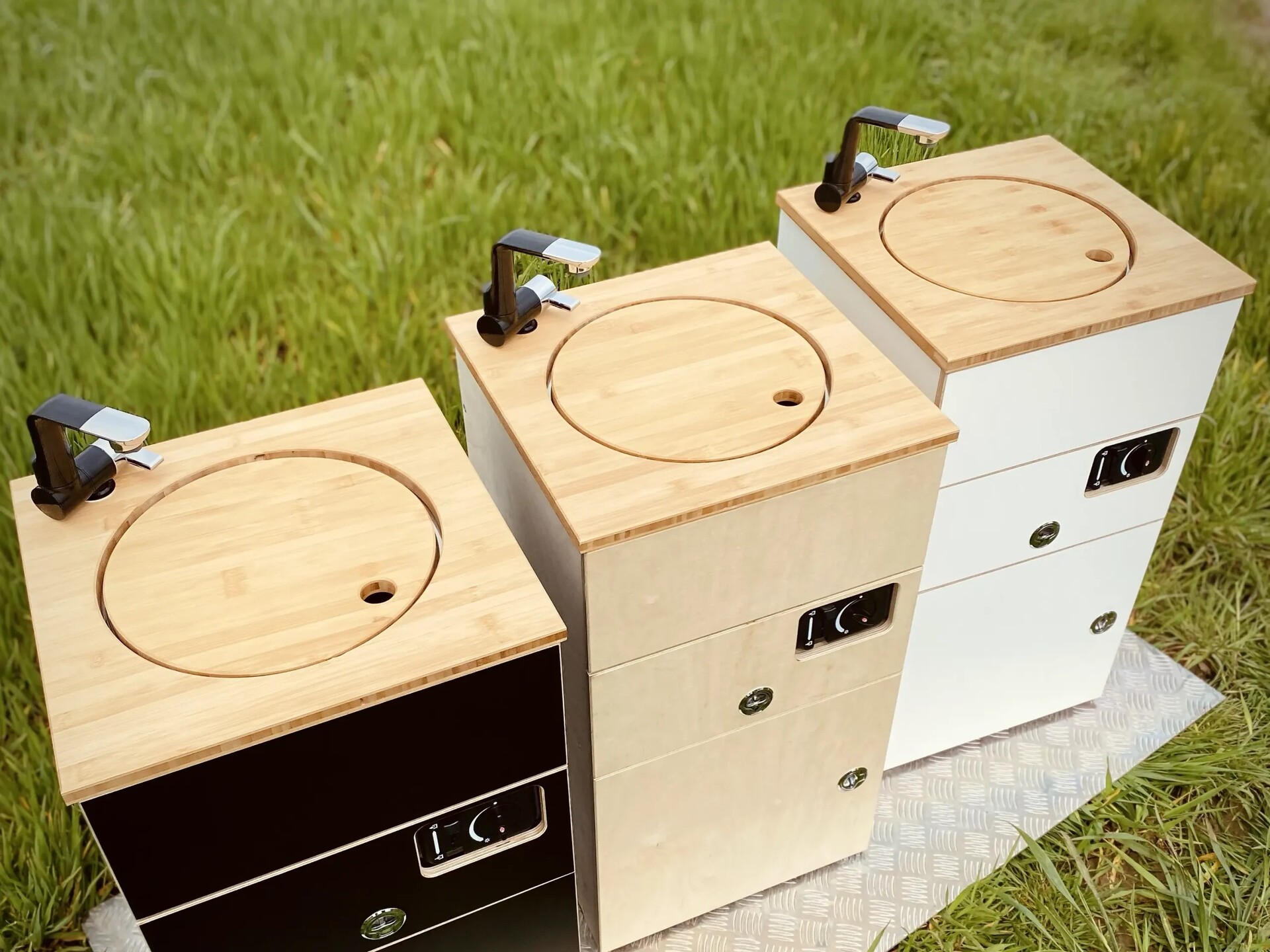 Tre stilfulde køkkenmoduler med bambusoverflader og vandhaner står på en eng og tilbyder en praktisk løsning til camping.