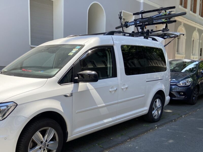 Ein weißer Transporter mit einem Fahrradträger auf dem Dach steht auf einer Straße, umgeben von weiteren Fahrzeugen, ideal für Campingausflüge und Outdoor-Aktivitäten.